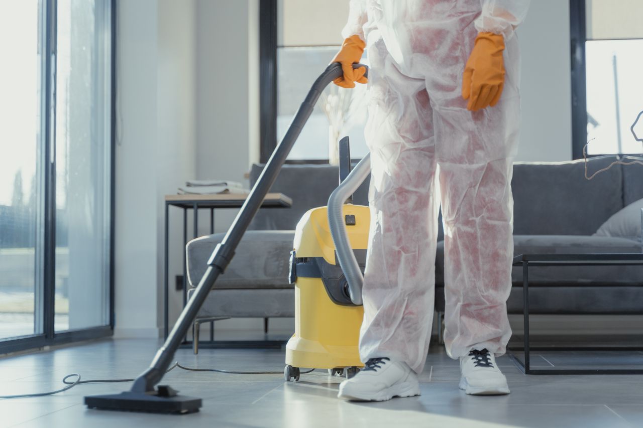 services-03 A cleaner in protective coveralls and gloves vacuuming a modern living room.